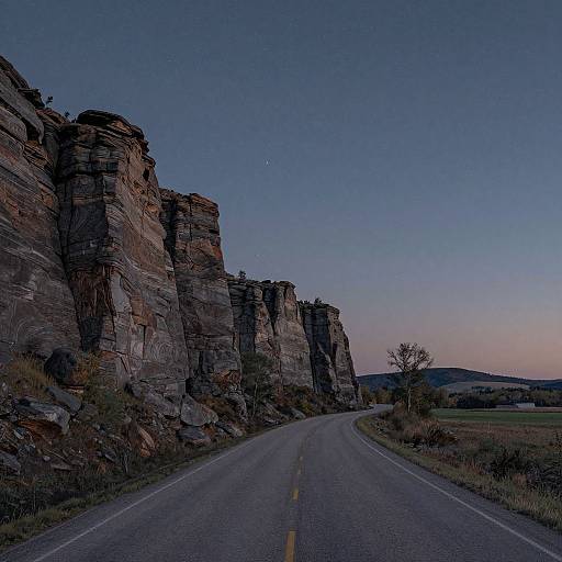 Photograph of a winding road at dusk, flanked by towering, rugged cliffs under a deep blue, twilight sky.