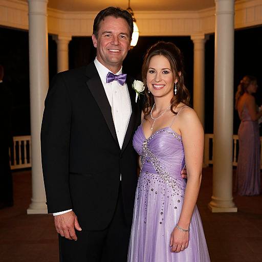 Photograph of a smiling couple at an evening event; man in black tuxedo and purple bowtie, woman in lavender strapless gown.