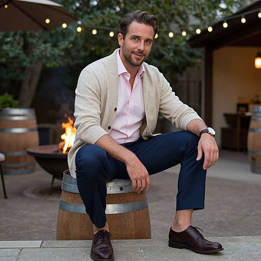 Photograph of a handsome bearded man with dark hair, wearing a white cardigan, pink shirt, and dark pants, sitting on a wooden barrel
