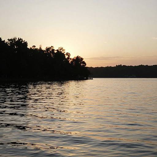 Photograph of a serene sunset over a calm lake, with silhouetted trees on the left and gentle ripples reflecting the orange and yellow sky