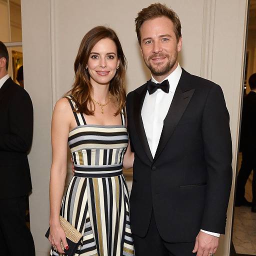 Photograph of a smiling, brown-haired woman in a striped dress and a bearded man in a black tuxedo at a formal event.