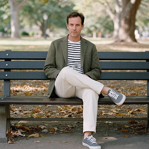 Photograph of a man with short brown hair, green blazer, white pants, black and white striped shirt, and black sneakers, sitting on a