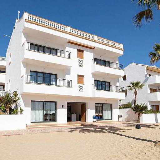 Photograph of a bright white, three-story beachfront apartment building with black-framed windows, balconies, and palm trees under a clear blue sky