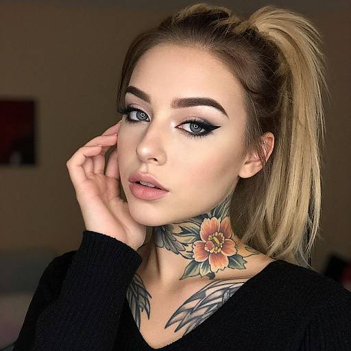 Photograph of a woman with blonde ponytail, black eyeliner, flower and feather tattoo on neck, hand near face, wearing black top, indoor