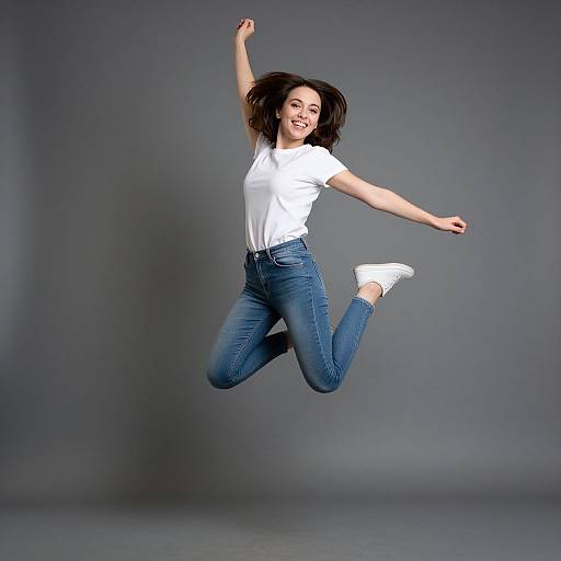 Photograph of a smiling woman with dark hair, wearing a white t-shirt and blue jeans, mid-air jumping against a gray background.