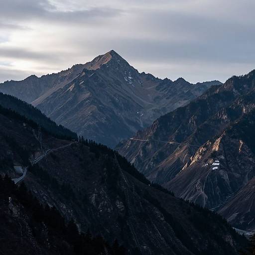Photograph of a mountain range at dusk, with dark, shadowed peaks in the foreground and a sunlit, rugged summit in the background under a