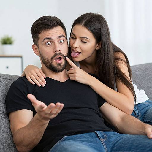 Photograph of a surprised bearded man in a black shirt, sitting on a gray couch, as a playful woman with long dark hair and white top