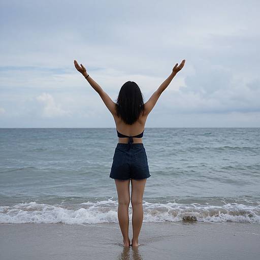 Photograph of a woman with long dark hair, wearing a black bikini top and denim skirt, standing on a beach with arms raised, facing the ocean
