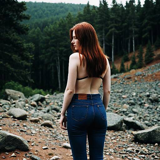 Photograph of a red-haired woman with fair skin, wearing a black bra and blue jeans, standing in a rocky forest clearing, looking over her shoulder