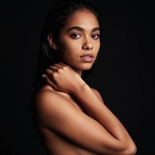 Photograph of a topless, dark-skinned woman with long black hair, brown eyes, and pink lips, against a black background, arms crossed