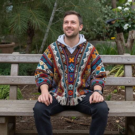Photograph of a smiling man with short brown hair, wearing a colorful, patterned sweater over a white hoodie, and black pants, sitting on a