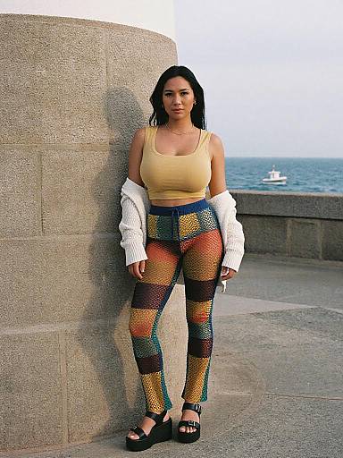 Photograph of a young woman with black hair, wearing a yellow tank top, colorful patterned leggings, white jacket, and black sandals, leaning against