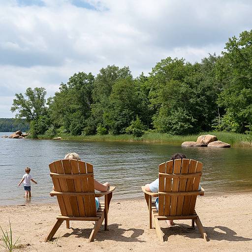 Photograph of two wooden Adirondack chairs on a sandy lake shore, with a child wading in the water, surrounded by lush green trees
