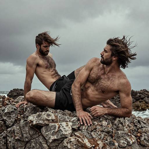 Photograph of two shirtless, bearded, muscular men with wind-swept hair, sitting on rocky shoreline in black shorts, under cloudy sky