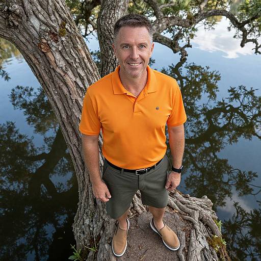 Photograph of a middle-aged man with short brown hair, wearing an orange polo shirt, dark green shorts, and tan shoes, standing against a tree