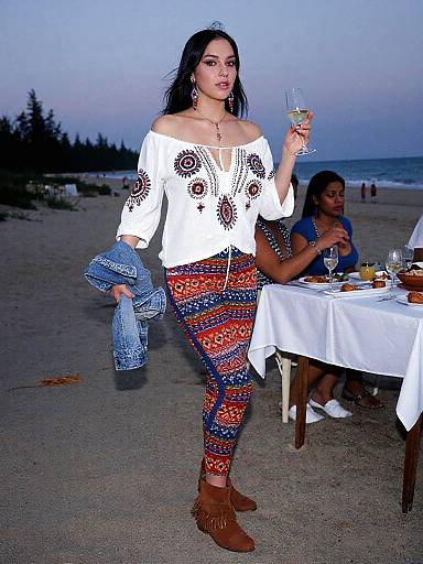 Photograph of a Latina woman with long black hair in a white off-shoulder top and colorful patterned pants, holding a glass of white wine
