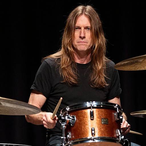 Photograph of a middle-aged man with long brown hair, wearing a black t-shirt, playing a brown wooden drum set on a dark stage.