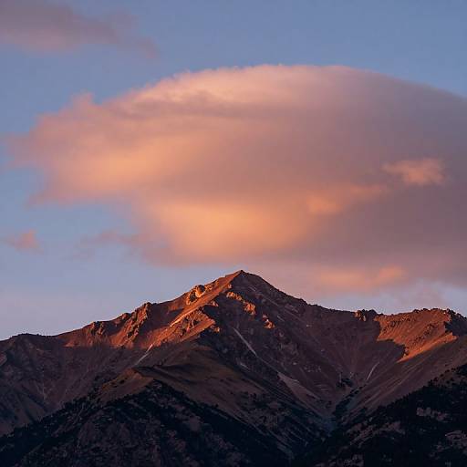 Photograph of a rugged mountain peak bathed in warm orange sunset light, with a large pink and orange cloud against a clear blue sky.