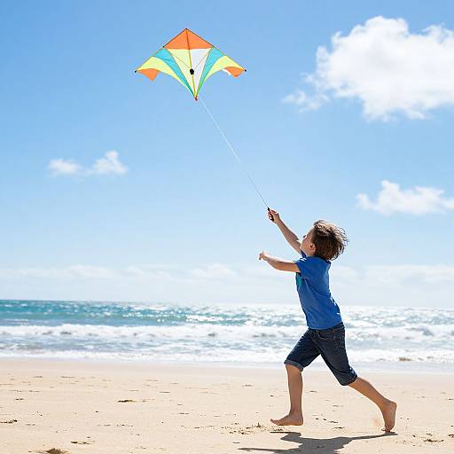 Photograph of a young boy with curly brown hair flying a colorful kite on a sunny beach with blue sky and ocean waves.