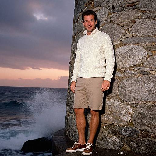 Photograph of a smiling man in a white sweater, beige shorts, and brown sneakers, standing against a stone wall by a crashing ocean wave at sunset