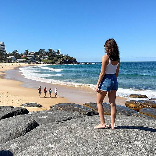 Photograph of a woman with long brown hair in a white tank top and blue denim shorts standing on rocky shore, facing beach with clear blue sky and