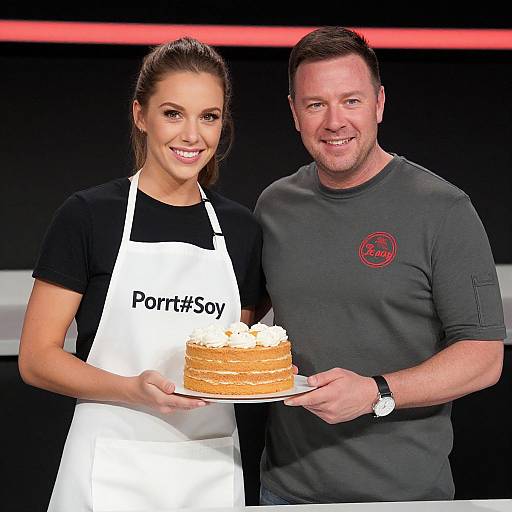 Photograph of a smiling woman in a black shirt and white apron, and a man in a gray shirt holding a cream-topped cake together on