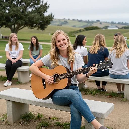 Photograph of a smiling blonde woman with long hair, wearing a white t-shirt and blue jeans, playing an acoustic guitar while seated on a concrete bench