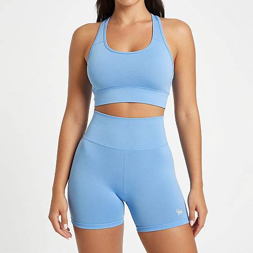 Photograph of a fit woman with tan skin wearing light blue sports bra and high-waisted shorts, white background, athletic attire.