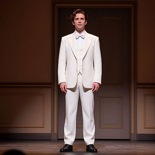 Photograph of a young man with fair skin and dark brown hair, standing in a dimly lit room wearing a white tuxedo, black shoes