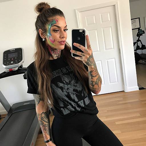 Tattooed woman with colorful face paint takes mirror selfie in gym room, wearing black t-shirt and black pants, wooden floor, treadmill in background