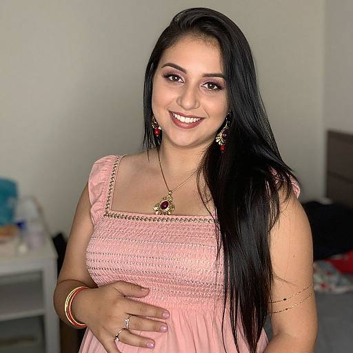 Photograph of a smiling woman with long black hair, wearing a pink dress, gold necklace, earrings, and red bangles, standing in a softly