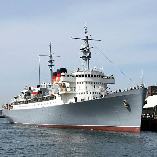 Photograph of a large white naval ship with a black and red smokestack, docked in calm blue water under a clear sky.