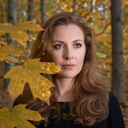 Photograph of a fair-skinned woman with long, wavy brown hair, wearing a black top, standing in a forest with autumn leaves, sunlight