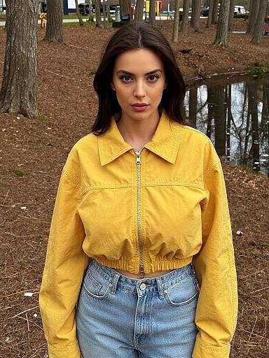Photograph of a young woman with dark hair, wearing a yellow jacket and blue jeans, standing in a wooded area with a small pond in the background