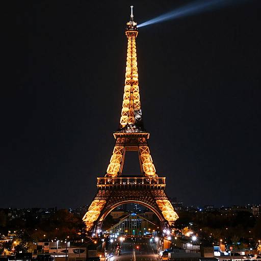 Photograph of the Eiffel Tower illuminated with bright yellow lights at night, with a blue laser beam pointing towards the top. Dark sky and city