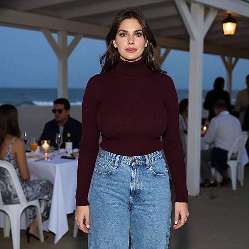 Photograph of a dark-haired woman in a black turtleneck and high-waisted blue jeans, standing in a beachside restaurant with sunset sky