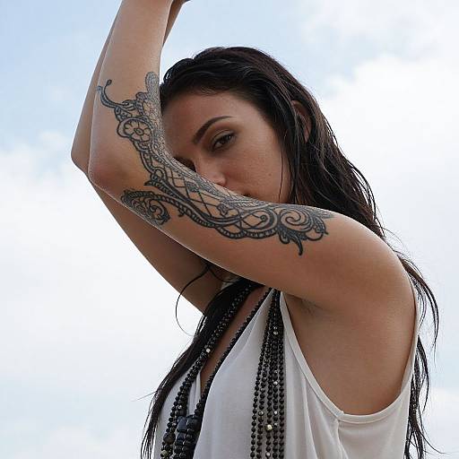 Photograph of a woman with long black hair, raising her arms to reveal an intricate black tattoo on her right arm. She wears a white sleeveless