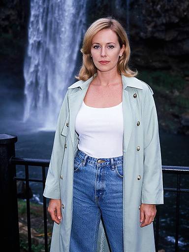 Photograph of a blonde woman with light skin, wearing a white tank top, blue jeans, and a light green coat, standing in front of a
