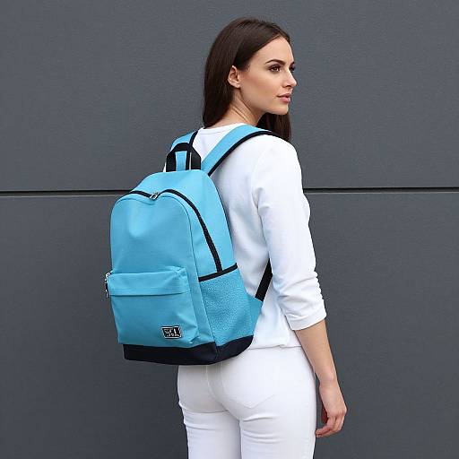 Photograph of a woman with long black hair, wearing a white long-sleeve shirt and white pants, with a light blue backpack, standing against
