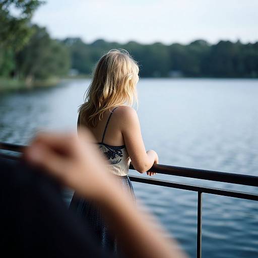 Photograph of a blonde woman in a black lace dress standing by a railing, gazing at a calm lake with trees in the background, blurred hand