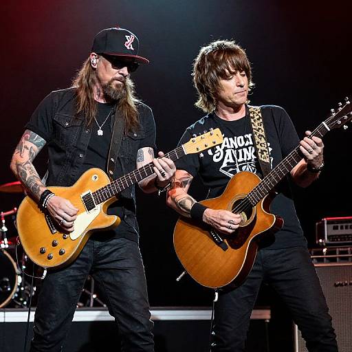 Photograph of two tattooed male guitarists on stage, wearing black clothing and hats, playing electric guitars with wooden bodies.