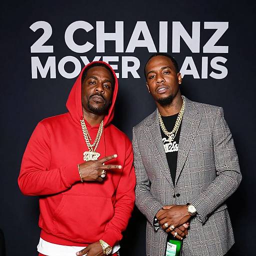 Photograph of two African-American men, one in a red hoodie and gold chains, the other in a gray checkered suit, standing against a black