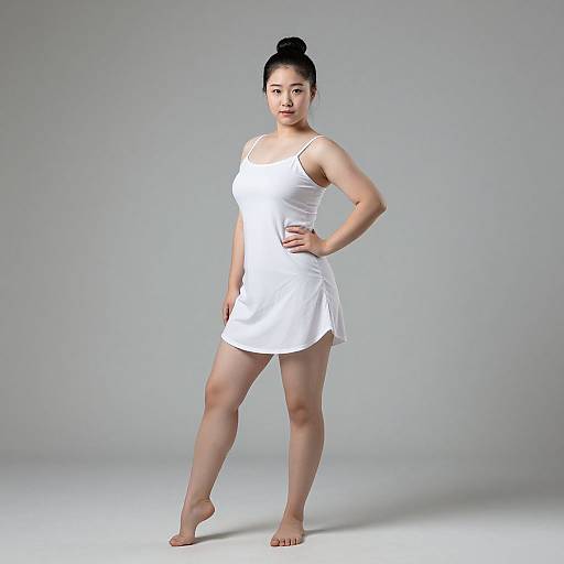 Photograph of an Asian woman with a bun, wearing a white, sleeveless, short dress, standing barefoot on a gray studio background.