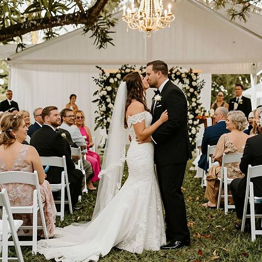 Photograph of a bride in a white lace off-shoulder gown and veil, and groom in a black suit, kissing at an outdoor wedding ceremony