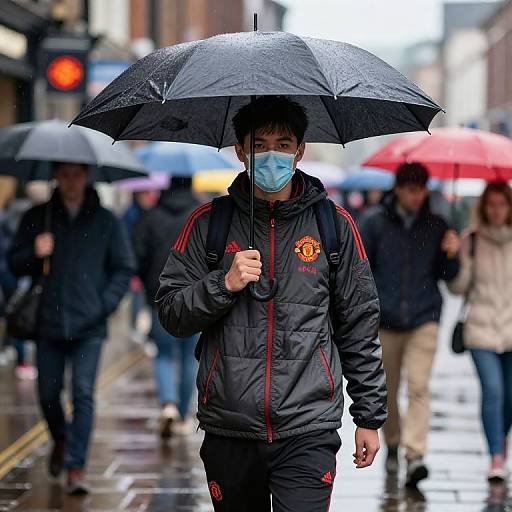 Photograph of an Asian man in a black tracksuit with red stripes, blue face mask, and black umbrella, walking in a rainy urban street with