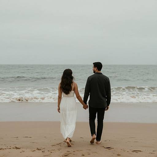 Photograph of a couple holding hands, walking on a sandy beach with gentle waves; woman in white dress, man in black suit.