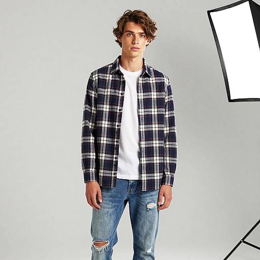 Photograph of a young man with wavy brown hair, wearing a black and white plaid shirt, white t-shirt, and ripped blue jeans,