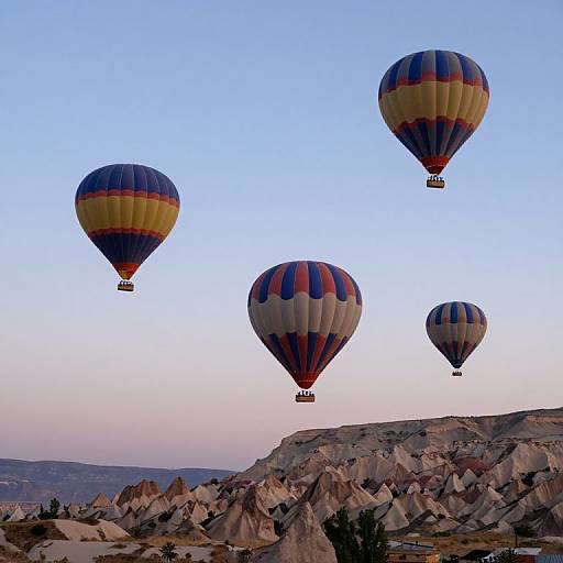 Photograph of four colorful hot air balloons floating over a rocky, mountainous landscape at sunrise, against a clear, blue sky.