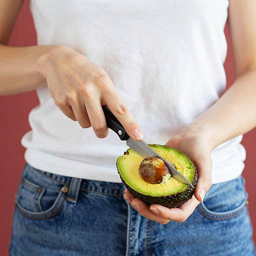 Photograph of a person in a white shirt and blue jeans, slicing a ripe avocado with a knife against a red background.