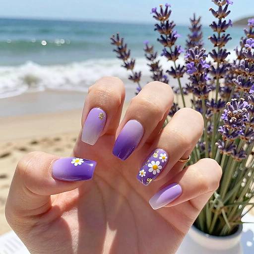 Photograph of a hand with long, purple-to-white gradient nails adorned with small daisy and gold floral stickers, holding lavender flowers, beach and ocean
